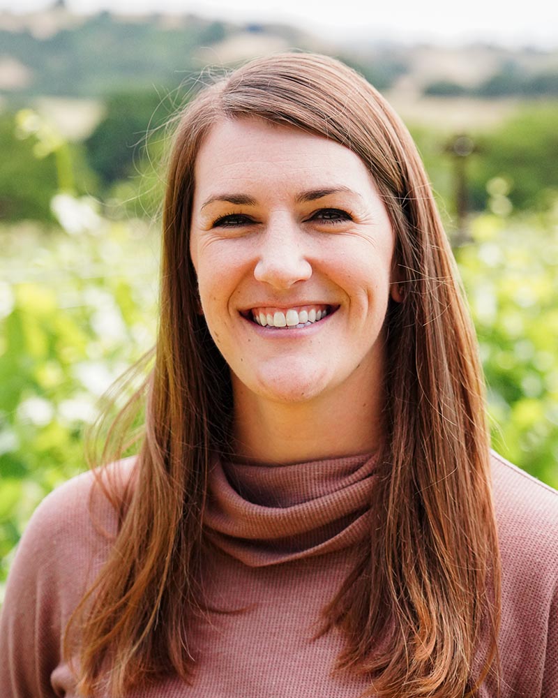 A smiling bio photo of Margaret Leonardi, with blurred green vines and hills in the background. 